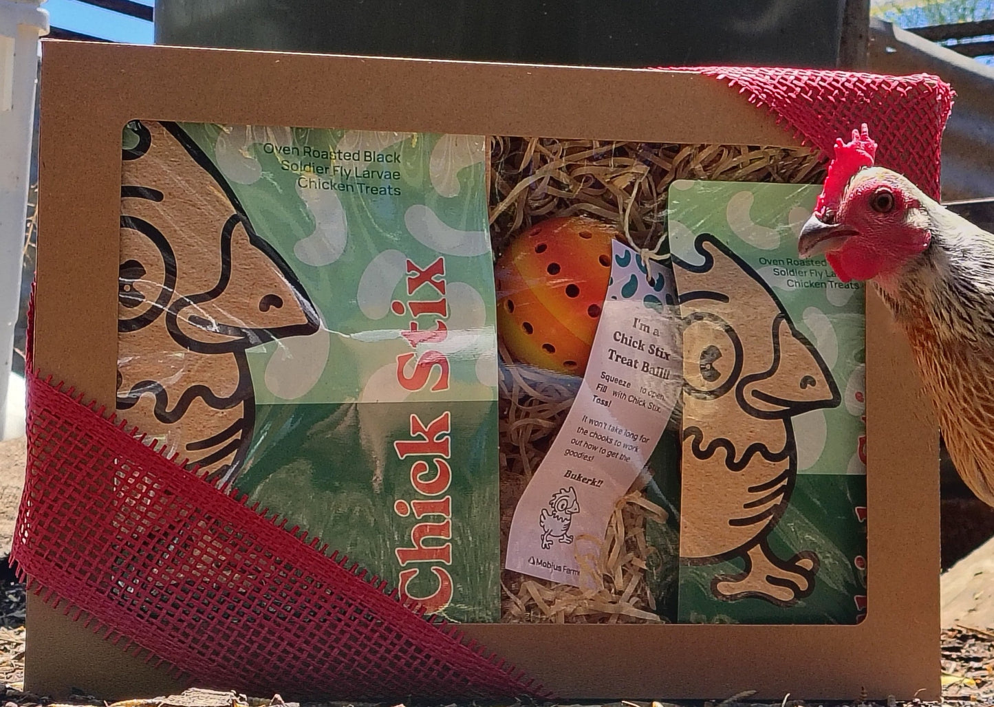 Box with a clear window has two bags of Chick Stix and a treat ball with a chicken peering over the edge